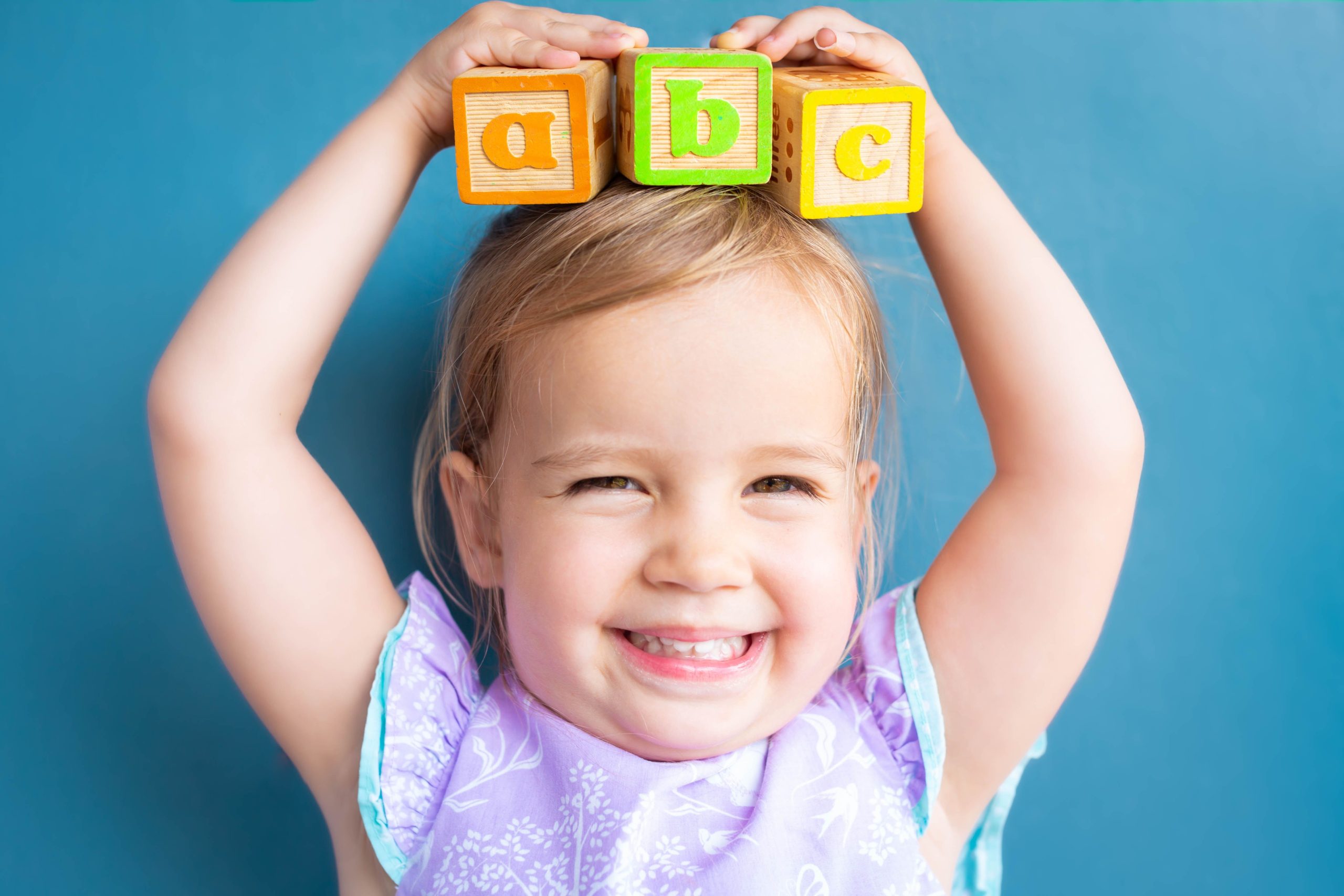 Small toddler in a purple shirt holding A B C blocks over their head.